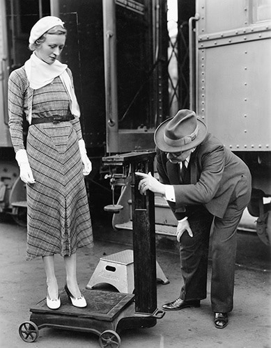 Vintage photo of man using obsolete scale to take woman's weight in front of a train