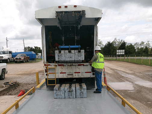 Michelli Weighing & Measurement service technician stands at the rear of a heavy duty test truck, using the remote to lower test weights onto a truck scale