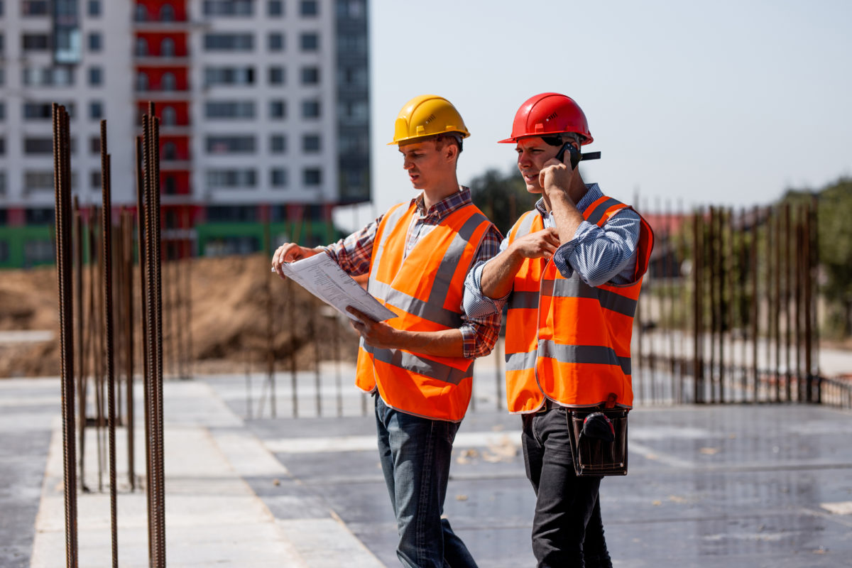 Two structural engineers dressed in shirts, orange work vests and helmets explore construction documentation and talk by phone on the building site near the steel frames