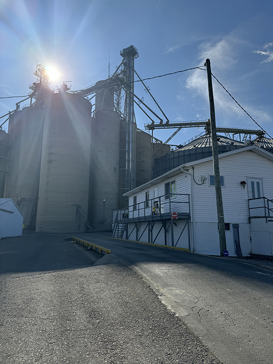 Image of a truck scale and white scale house with industrial silos in the background